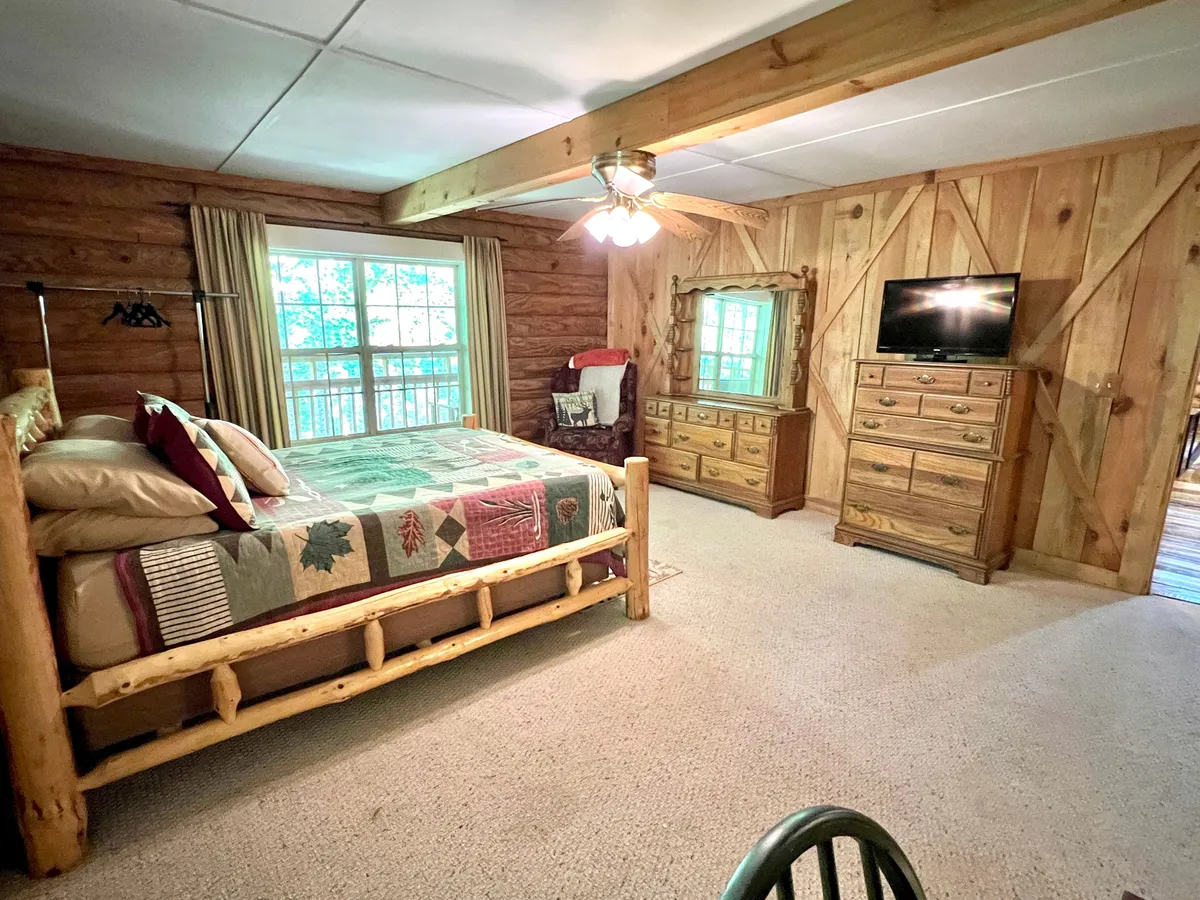 A cozy master bedroom with a rustic log bed topped with a quilted bedspread, a dressing table, TV, and a window letting in soft natural light. The warm wooden accents and thoughtful decor create an inviting atmosphere ideal for relaxation.