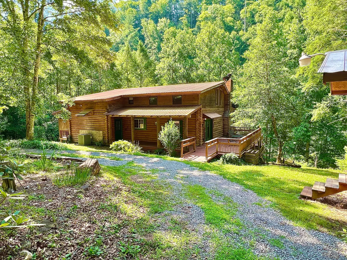 A spacious log cabin nestled among lush greenery, featuring a large deck area perfect for outdoor gatherings. The surrounding trees enhance the sense of seclusion and connection with nature, while the open deck is ideal for relaxing or socializing.
