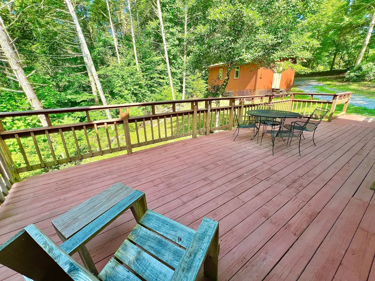 A spacious wooden deck with a black metal table and chairs, surrounded by lush greenery and an orange cabin visible in the background. The deck offers a perfect space for outdoor dining, relaxing with a book, or gathering with family and friends under the open sky.