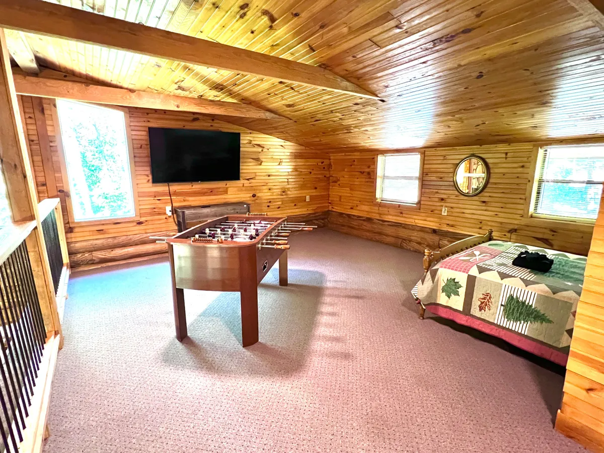The image captures a playful yet relaxed loft space featuring a foosball table, a cozy bed with a patterned quilt, a flat-screen TV on the wall, and warm wooden walls. Natural light filters in, creating a welcoming atmosphere ideal for both play and relaxation.