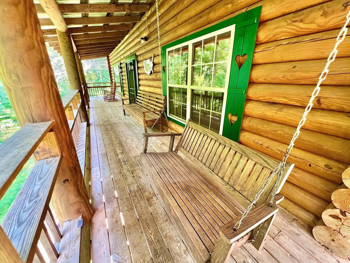A charming porch with wooden swing benches, green window frames, and classic log cabin walls, inviting guests to unwind and enjoy the outdoors. The space offers plenty of room for socializing or peaceful solitude, surrounded by nature.