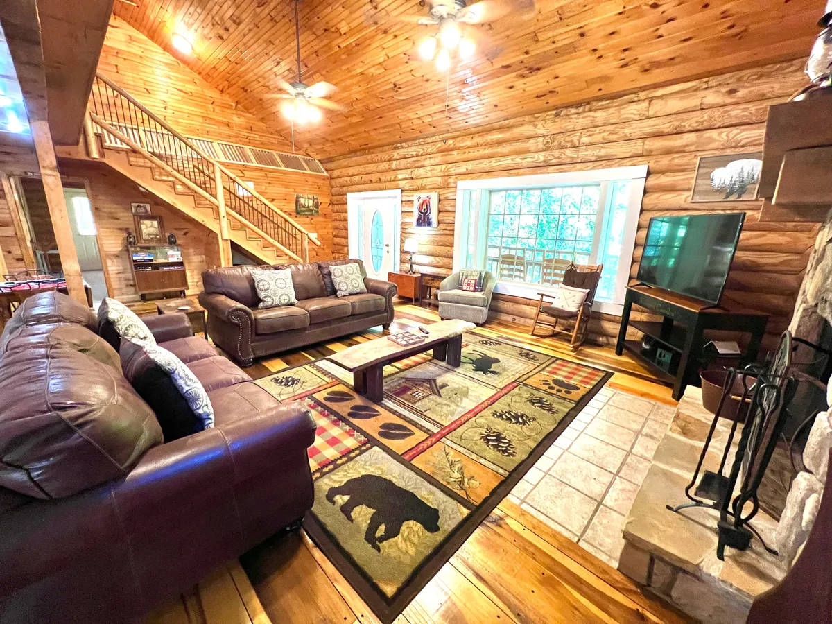 A wide shot of the living room highlights several plush leather sofas arranged for group conversation, a rustic fireplace, a large window drawing in natural light and forest scenery, and a wooden staircase that adds to the cabin's character. The setting suggests moments of sharing warm drinks, movie nights, and family celebrations.