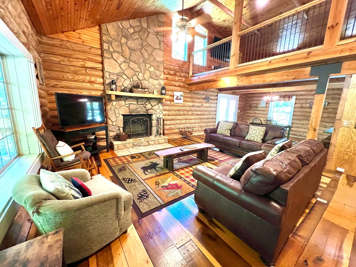 This angle of the living area showcases the stone fireplace as a focal point, surrounded by leather sofas and a wildlife-themed rug. Large windows bathe the space in natural light, while the layout encourages group interaction for family gatherings, game nights, and quiet evenings by the fire.