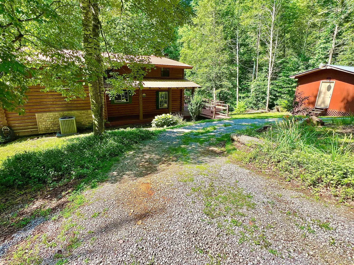 A full view of the log cabin nestled among tall trees, with a gravel driveway and a red shed nearby. The setting is quiet and surrounded by woods, evoking a true sense of seclusion and the promise of outdoor fun.