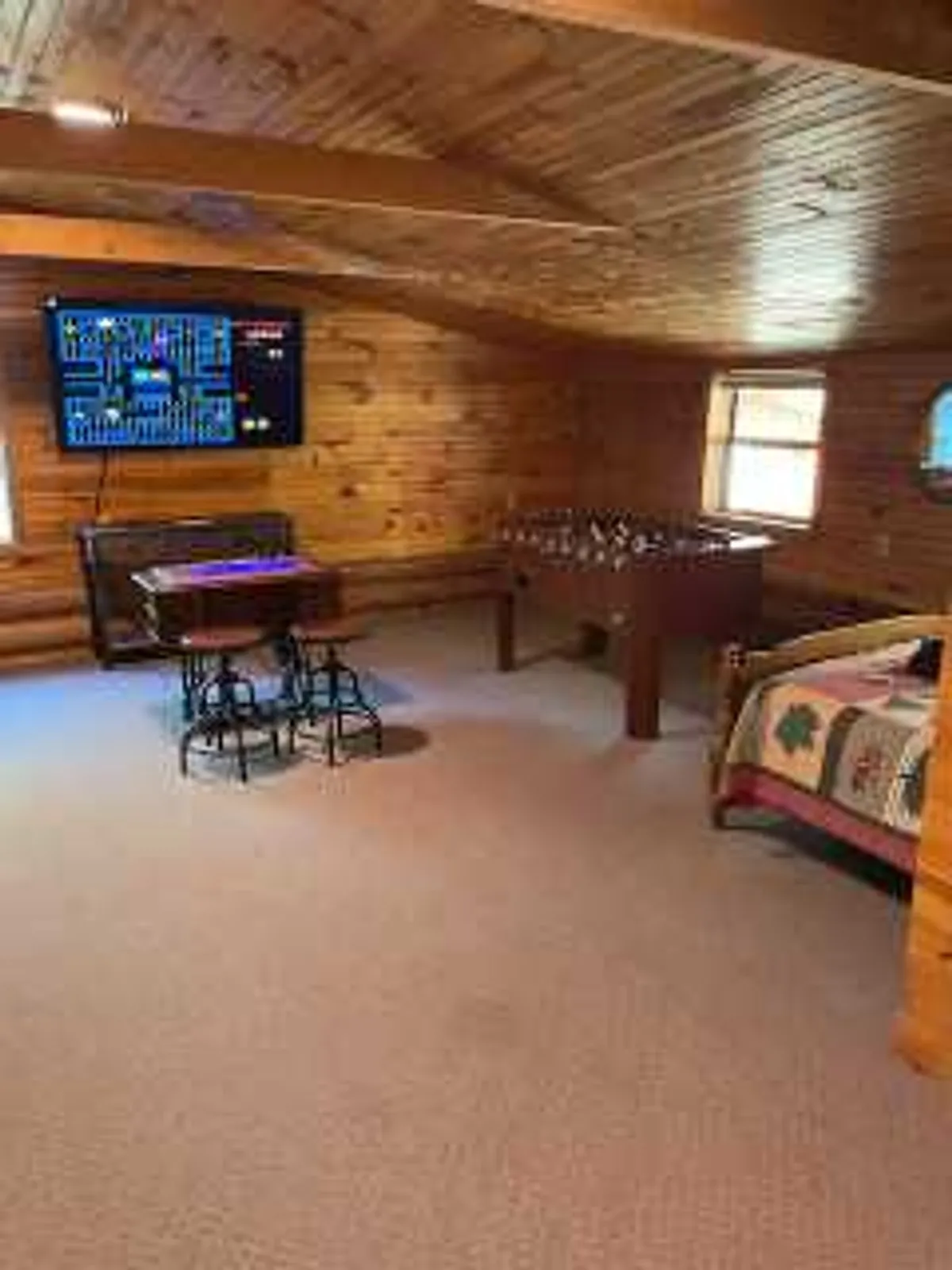 A lively scene featuring a foosball table, a bed with a patterned quilt, a small table with chairs, and a television displaying an arcade game. The overall mood is energetic and fun, ideal for gatherings, late-night tournaments, or quiet moments on the bed.