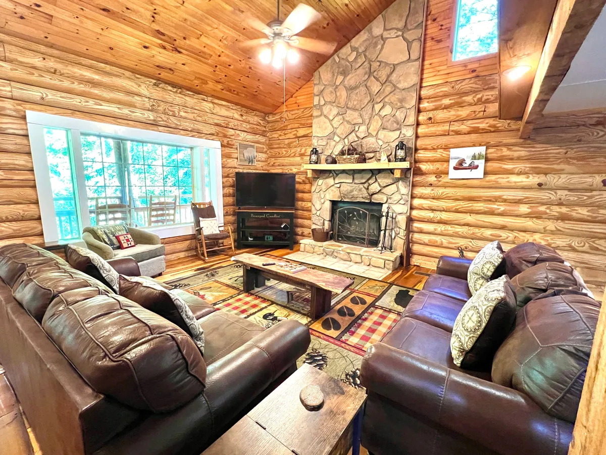 This image captures a spacious living area featuring a prominent stone fireplace, large windows that let in abundant sunlight and views of the forest, plush leather sofas arranged for group gatherings, and a patterned rug that adds rustic charm. The atmosphere is cozy and welcoming, perfect for family gatherings or quiet relaxation by the fire.