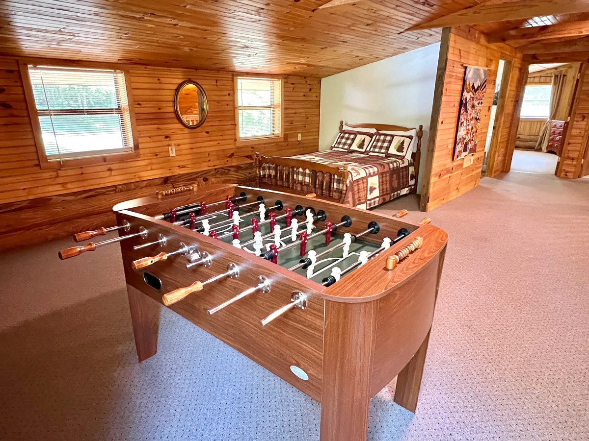 This view highlights the game room loft with a foosball table set in front of a bed covered with a vibrant quilt. Large windows let in plenty of natural light, and the wooden walls maintain a rustic, welcoming charm. The layout suggests easy transition between playing games and relaxing in bed.