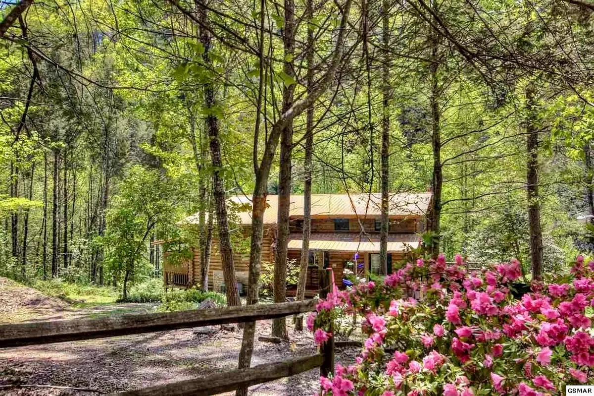A classic log cabin is framed by vibrant pink flowers and lush greenery, with a rustic wooden fence marking the edge of the property. The scene evokes a sense of welcoming natural beauty and outdoor enjoyment, suggesting opportunities for family gatherings, relaxation, and exploration just outside the cabin door.