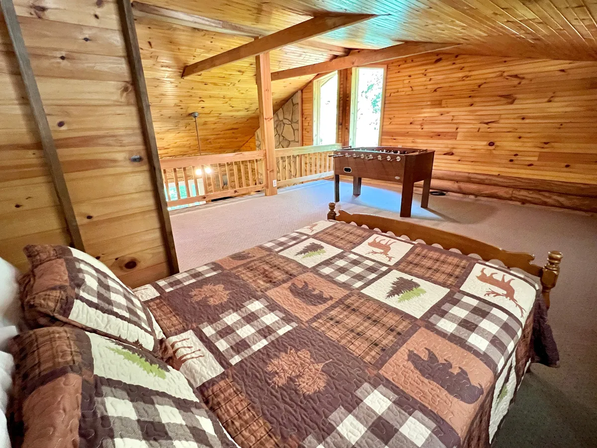 A rustic loft scene centered on a bed with a patterned quilt, a foosball table positioned for play, wooden walls and beams, and large windows letting in ample natural light. The space looks inviting for both resting and playing, with the windows hinting at beautiful outside views.