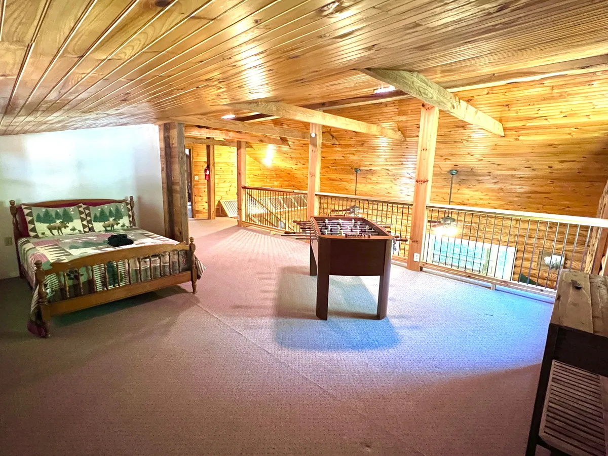 This image reveals a loft area featuring a full-sized bed with patterned bedding, a foosball table, wooden railing, and exposed ceiling beams. The bed is situated in a semi-private nook, while the foosball table is ready for games, clearly conveying the dual-purpose nature of the space. Warm wood tones create a playful yet cozy atmosphere.