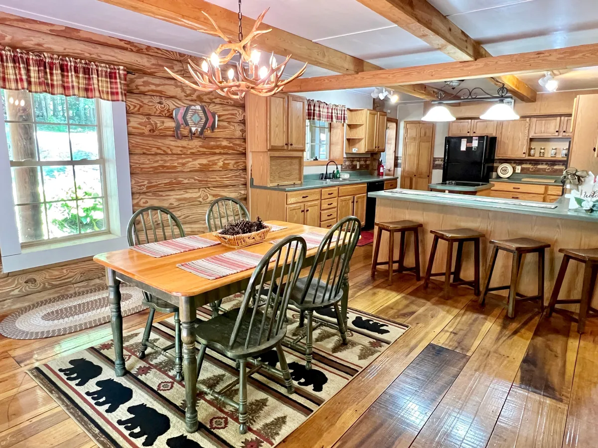 A rustic dining area featuring a large wooden table, antler chandelier, bear-themed rug, and a view into the adjoining kitchen bar area. The scene is set for family meals, celebrations, and lingering conversations.