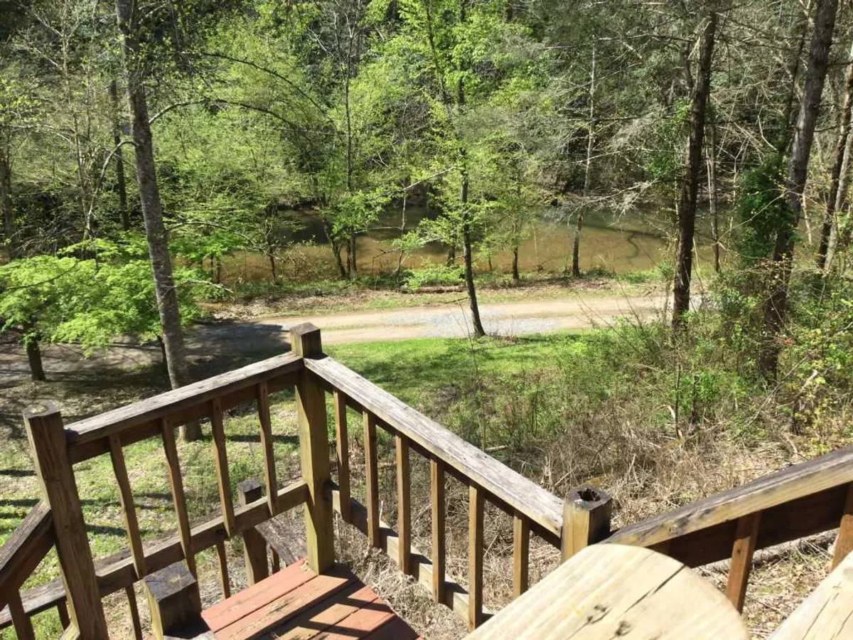 A wooden deck railing overlooks a canopy of lush greenery and calm water, with a dirt road nearby. The peaceful scene hints at opportunities for nature walks, bird watching, and tranquil moments spent outdoors.