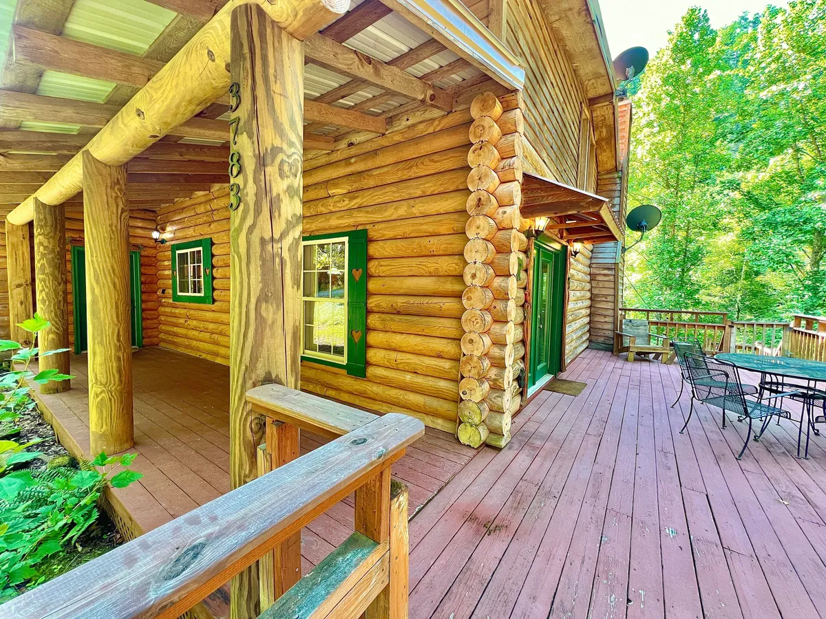 A log cabin structure with inviting green window shutters and a spacious deck area furnished for relaxation. The deck is surrounded by lush trees, creating a seamless connection to nature and offering a peaceful outdoor setting.
