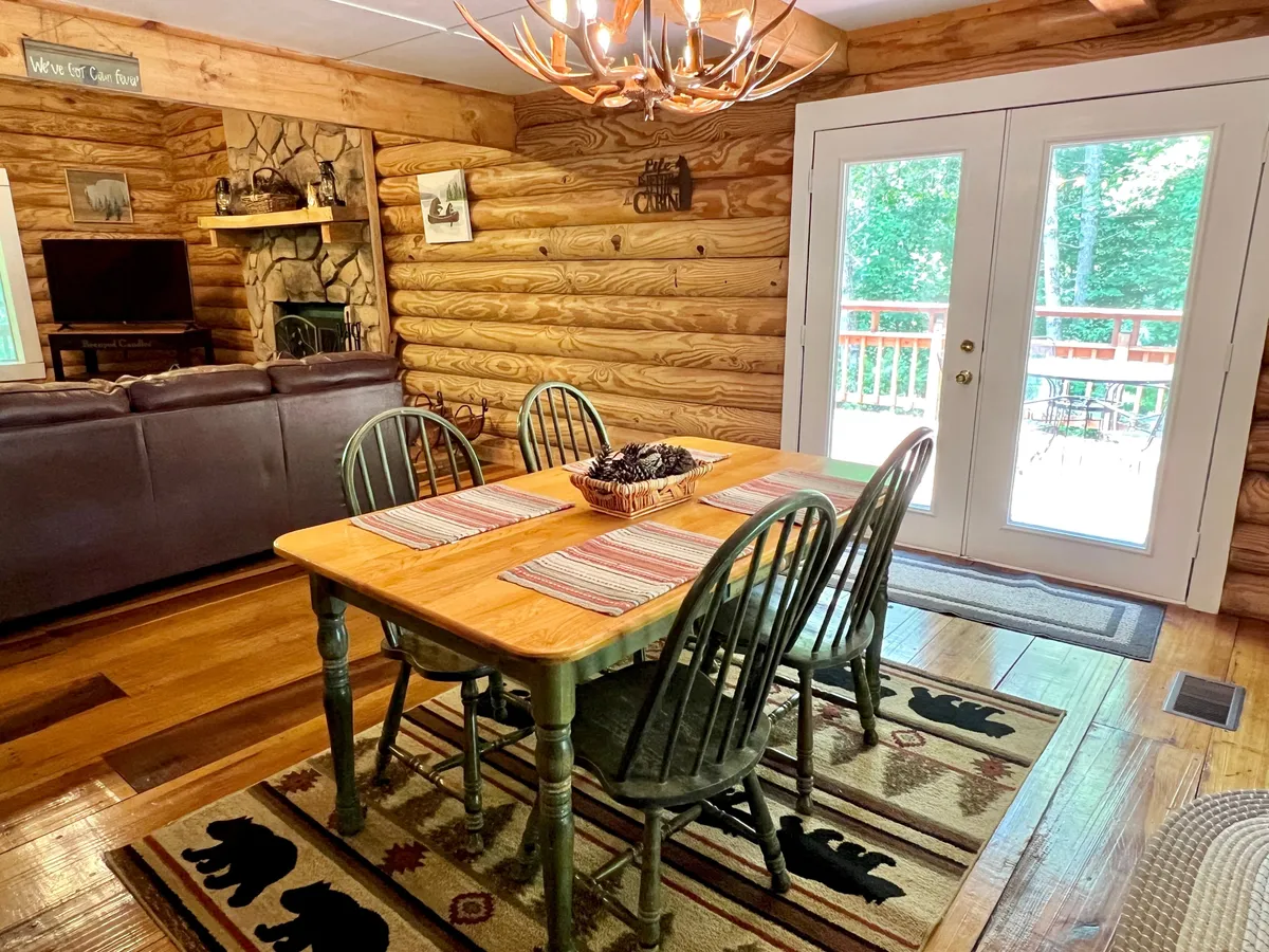 A warm, homey dining space with a wooden dining table, chandelier, log walls, a fireplace, and expansive windows. The setting is ideal for family dinners, game nights, and cozy gatherings, filled with laughter and shared moments.
