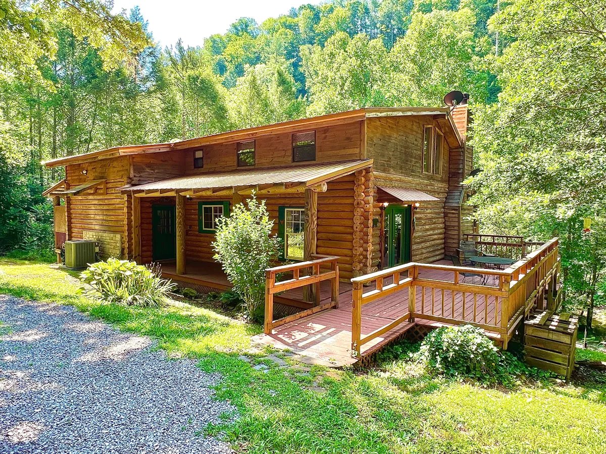 A cozy log cabin structure with a deck area set amidst abundant greenery and a gravel driveway. The inviting deck is ideal for gatherings, while the surrounding landscape provides plenty of space for exploration and relaxation in total privacy.